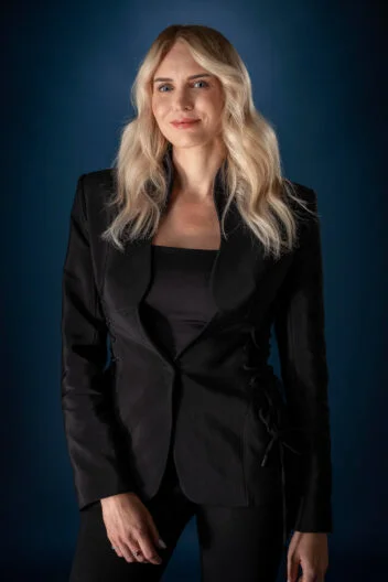 Portrait of a blonde woman in a black tailored suit standing against a dark blue studio background.