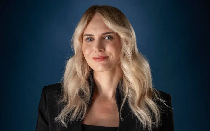 Portrait of a blonde woman in a black tailored suit standing against a dark blue studio background.