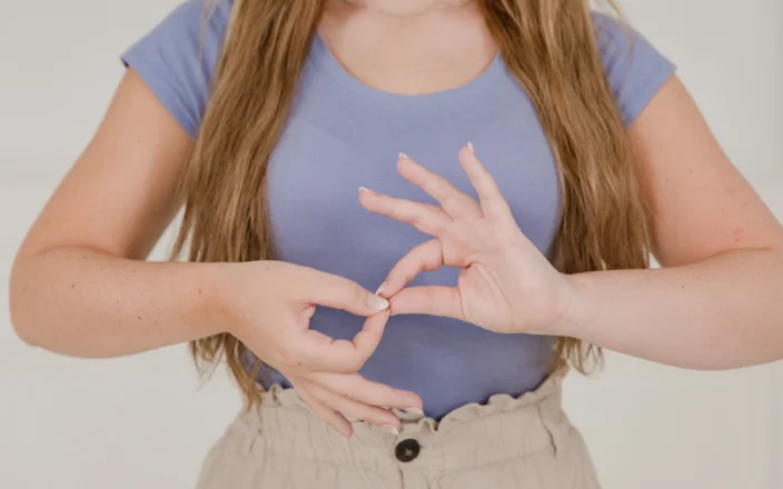 Woman demonstrating a hand gesture in sign language while standing against a neutral background.